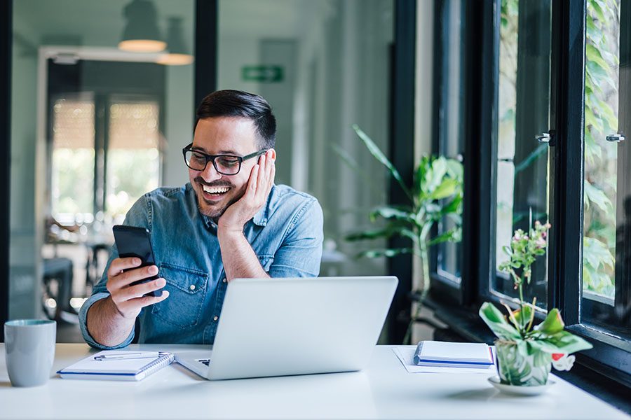 Business Insurance - Portrait of a Cheerful Young Business Owner Working on a Laptop ini a Modern Office While Holding a Phone in His Hand